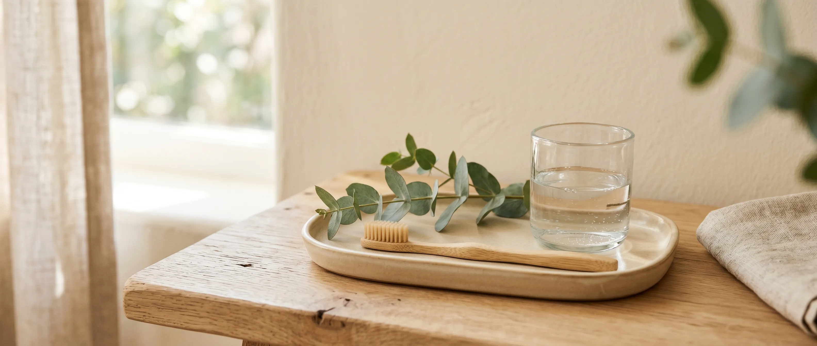 Minimalist oral care arrangement on a wooden tray with eucalyptus and a glass of water in soft natural light